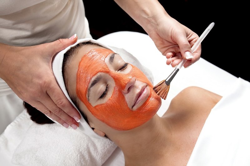 A carrot mask is applied to a girl’s face with a brush